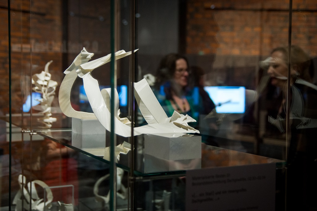 In einer gläsernen Vitrine stehen zerbrechliche Skulpturen, die aus weißen Bändern gebildet werden. Im Hintergrund Museumsbesucher.